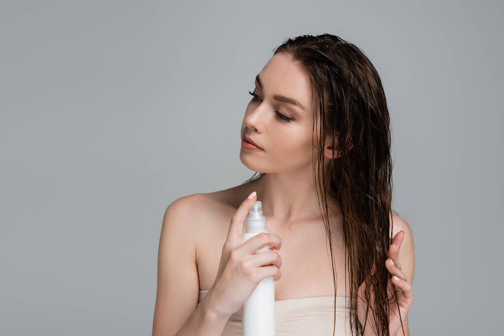bonita mujer joven con hombros desnudos aplicando producto de tratamiento en el cabello húmedo aislado en gris  - Foto, imagen