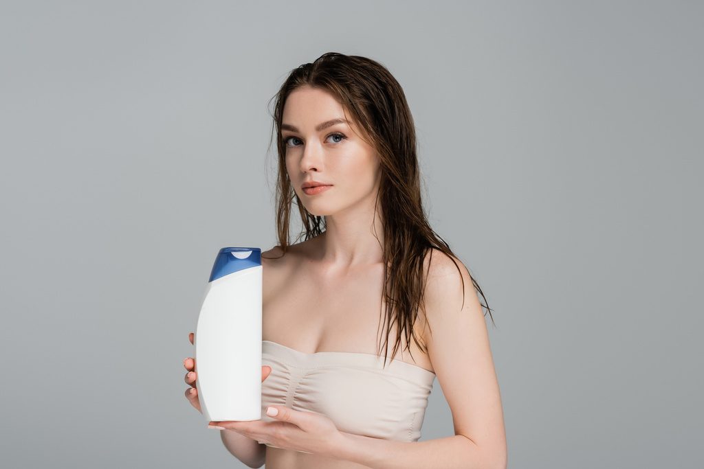 pretty woman with wet hair and bare shoulders holding shampoo bottle isolated on grey  - Photo, Image