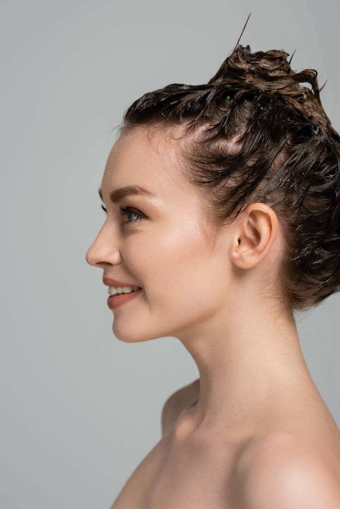 Радостная молодая женщина с пеной на голове смотрит в сторону, в то время как мытье волос изолированы на сером  - Фото, изображение