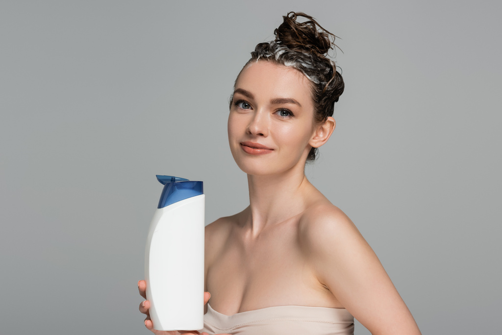 весела молода жінка з мокрим пінистим волоссям тримає пляшку з шампунем ізольовано на сірому
  - Фото, зображення