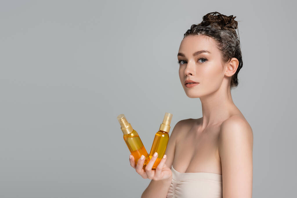 jovem morena com cabelo espumoso molhado segurando garrafas com óleo de tratamento isolado em cinza  - Foto, Imagem