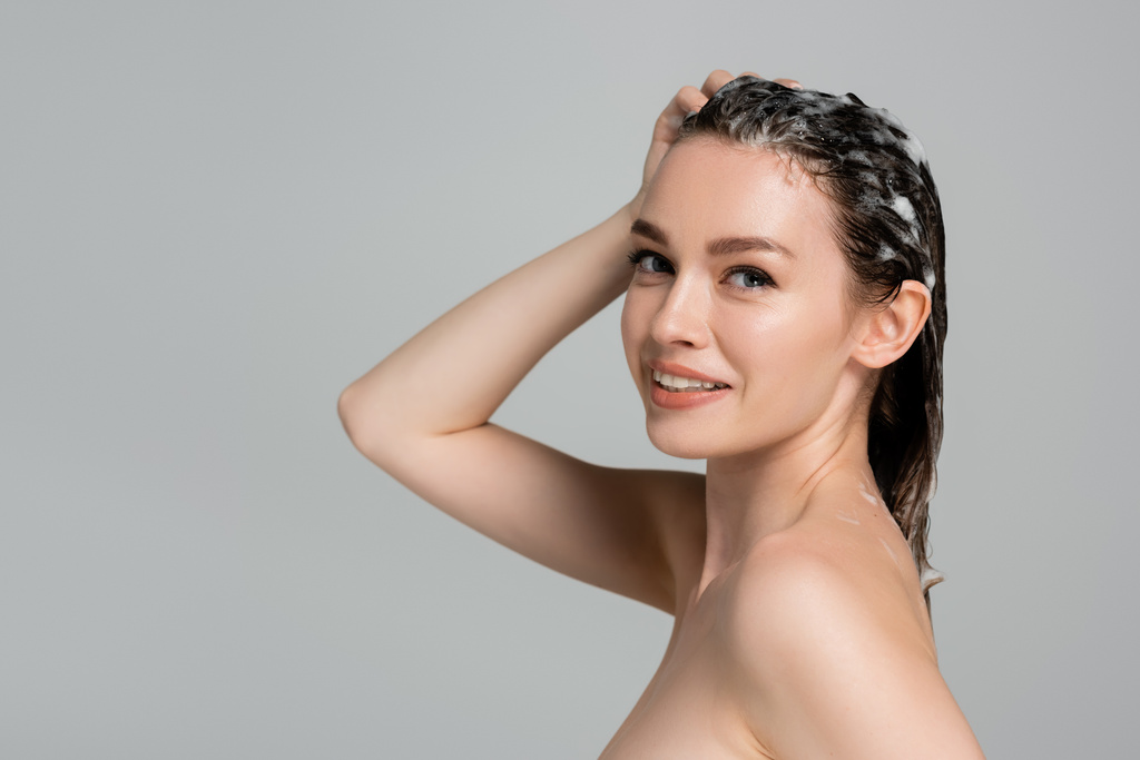 radosna młoda kobieta myjąca mokre i piankowe włosy odizolowane na szaro  - Zdjęcie, obraz