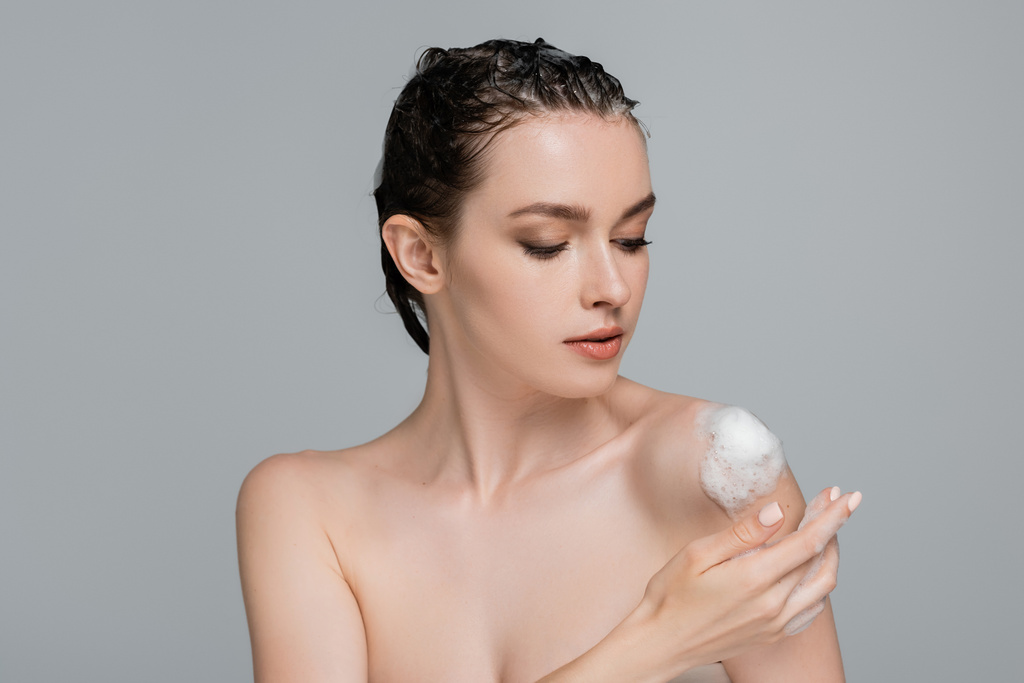 młoda kobieta z mokrymi włosami dotykająca gołego ramienia z białą pianką odizolowaną na szaro  - Zdjęcie, obraz