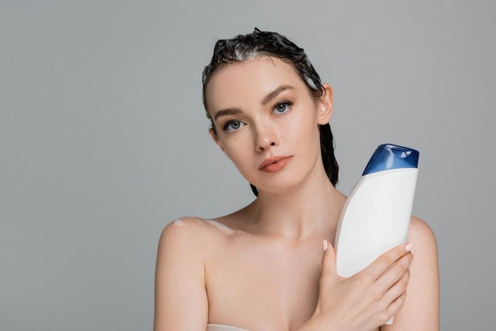 brunetka młoda kobieta z mokre piankowe włosy trzymając butelkę z szamponem izolowane na szary  - Zdjęcie, obraz