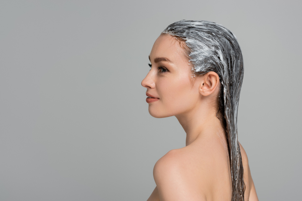 side view of cheerful young woman with bare shoulders and hair mask isolated on grey  - Photo, Image