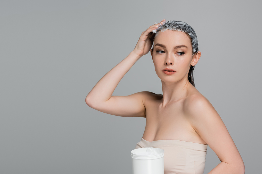 jeune femme aux épaules nues appliquant un masque sur cheveux mouillés et tenant un bocal isolé sur gris  - Photo, image