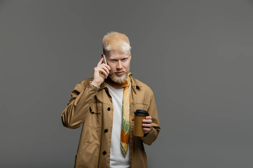 hombre albino barbudo en camisa chaqueta sosteniendo taza de papel y hablando en teléfono inteligente aislado en gris - Foto, Imagen