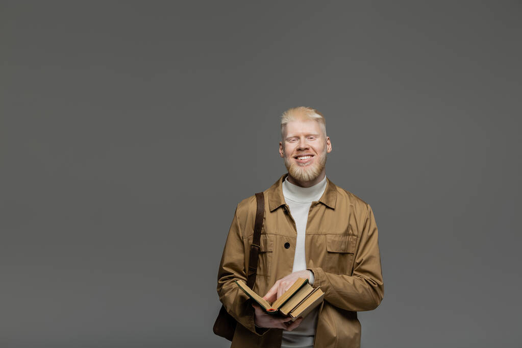 glücklicher Albino-Student mit Rucksack, der Bücher isoliert auf grau hält - Foto, Bild