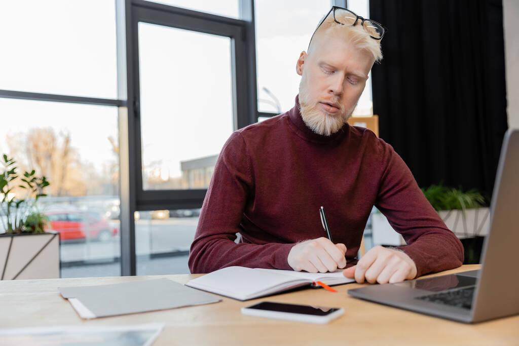 bearded albino businessman writing on notebook near devices on desk  - Photo, Image