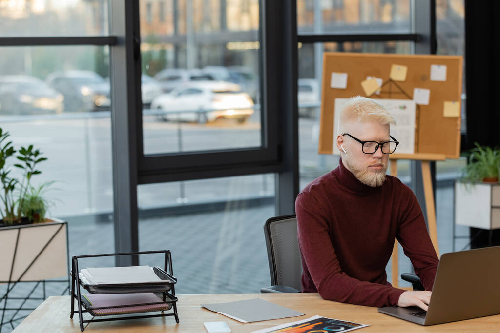 bebaarde albino zakenman in draadloze oortelefoon met behulp van laptop tijdens het werken in het kantoor  - Foto, afbeelding