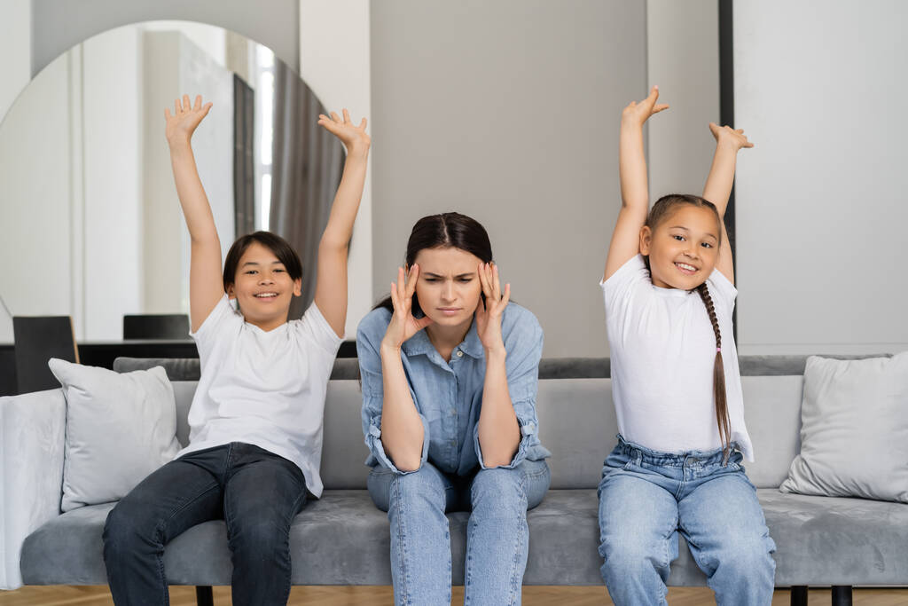 Cheerful asian kids sitting with raised hands near tired mother on couch at home  - Photo, Image