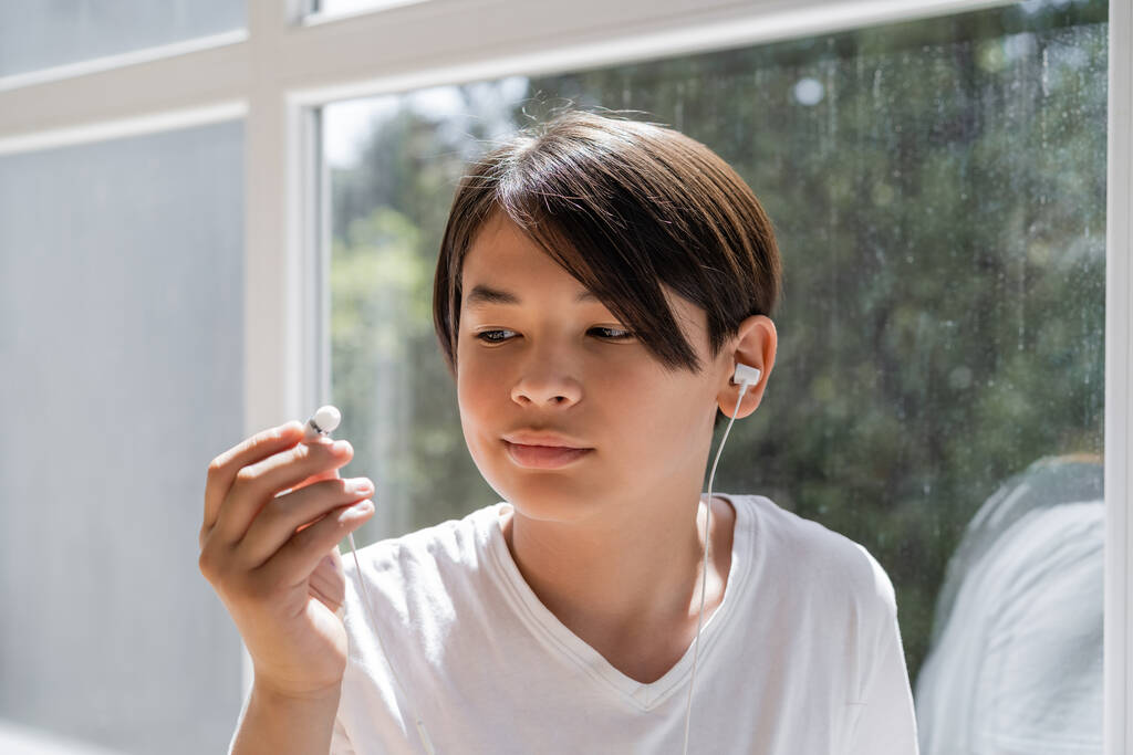 Niño asiático mirando auriculares con cable cerca de la ventana en casa  - Foto, imagen