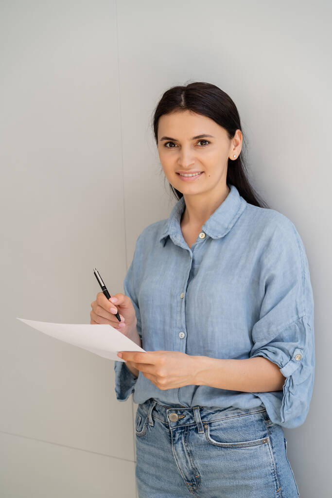 Iloinen nainen pitää kynää ja paperia lähellä seinää kotona  - Valokuva, kuva