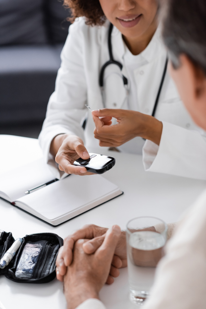 visão recortada do médico afro-americano em casaco branco segurando dispositivo de glicosímetro e tira de teste perto de paciente de meia-idade - Foto, Imagem