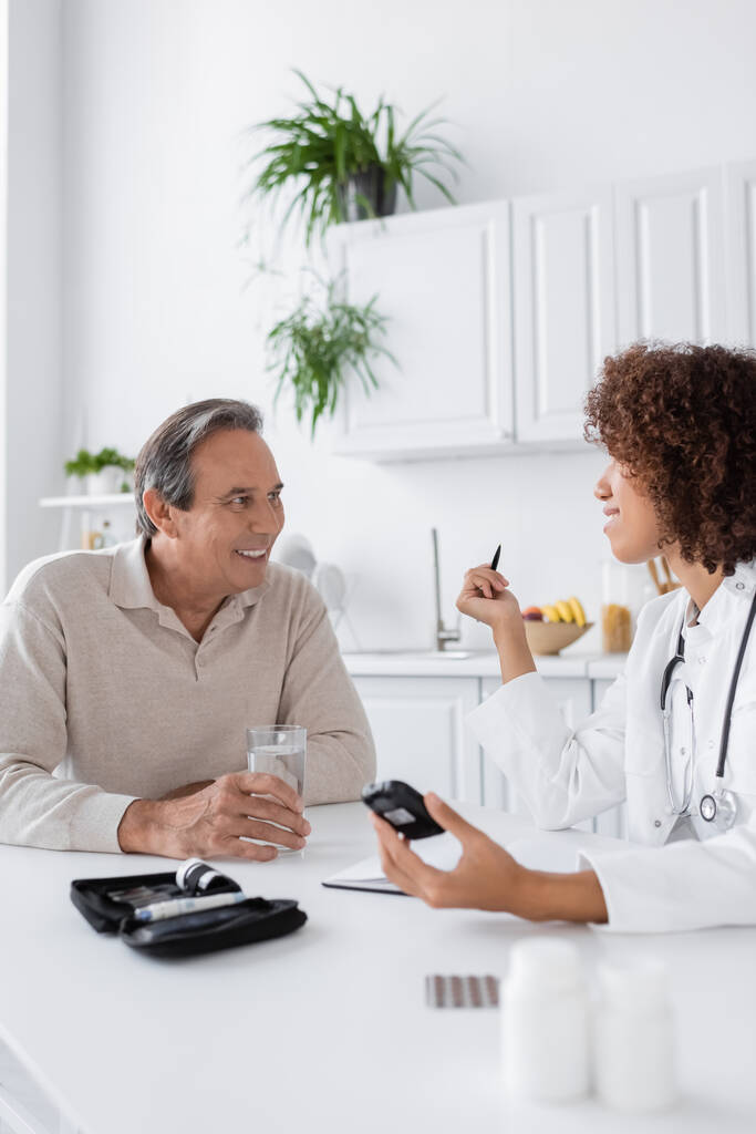 curly african american doctor holding glucometer near smiling middle aged patient  - Photo, Image
