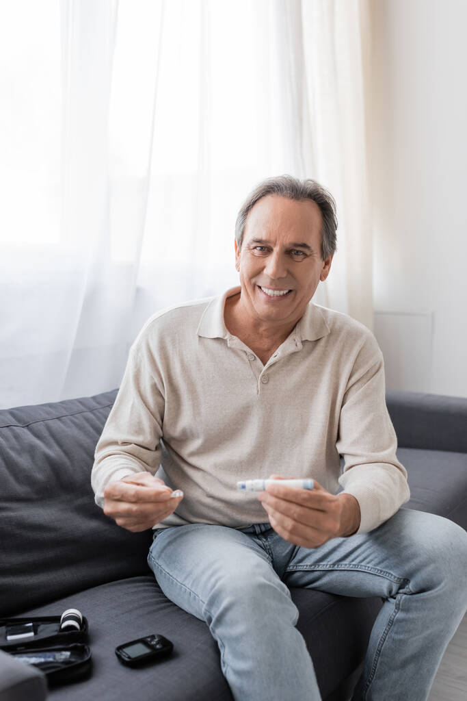 homem maduro alegre com diabetes segurando caneta lancet e tira de teste enquanto sentado no sofá na sala de estar  - Foto, Imagem