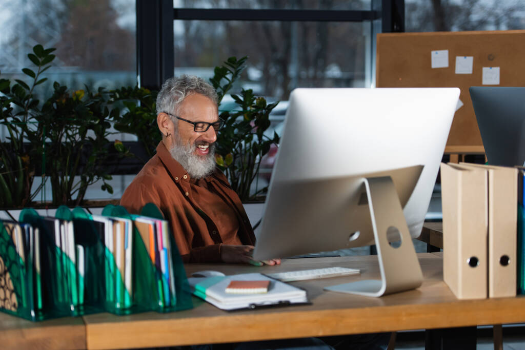 Veselý šedovlasý podnikatel pracující poblíž počítače a kancelářských papírů  - Fotografie, Obrázek