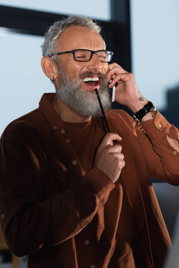 Allegro uomo d'affari barbuto con gli occhiali che tiene la matita mentre parla sullo smartphone in ufficio la sera  - Foto, immagini