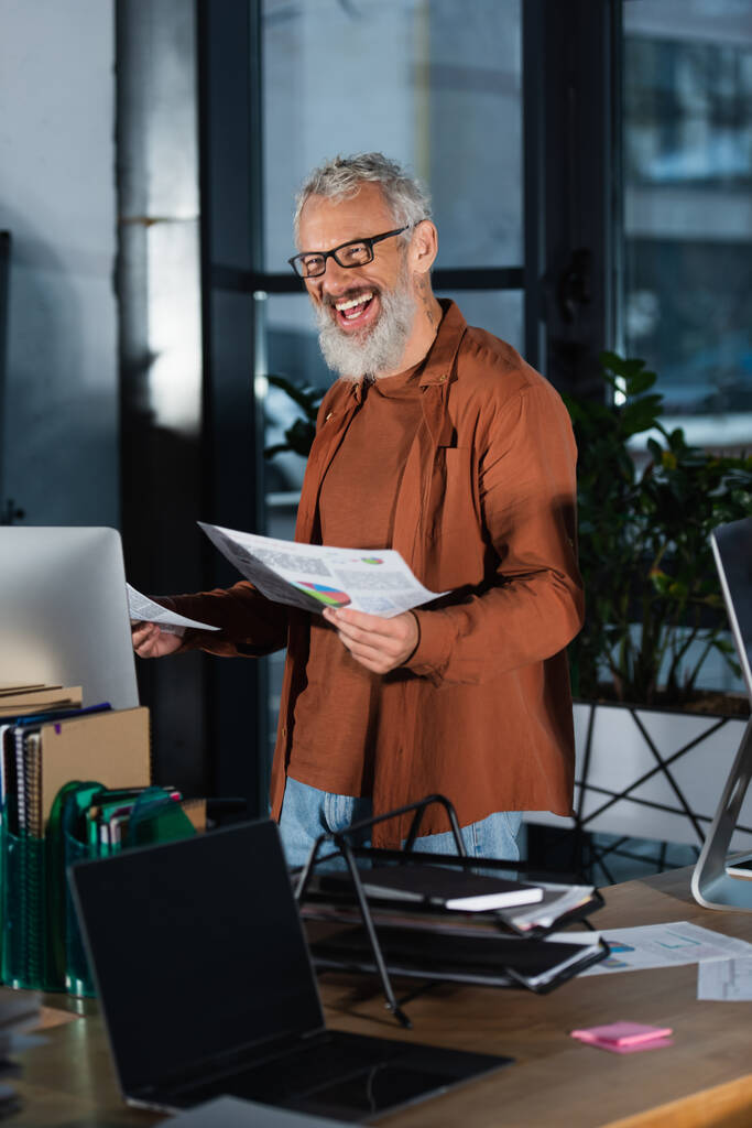 Веселий бізнесмен в окулярах тримає папери з графіками біля комп'ютера в офісі ввечері
  - Фото, зображення