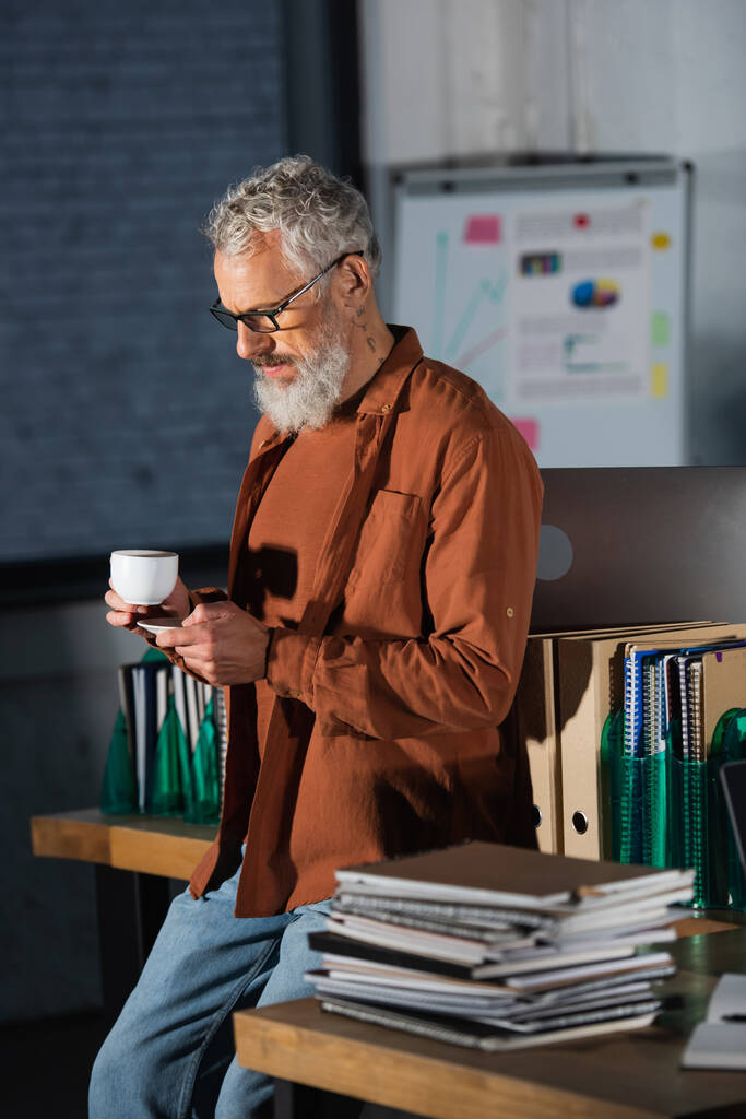 середнього віку бородатий бізнесмен з чашкою кави та смартфоном, що стоїть поруч з папками та блокнотами в офісі
 - Фото, зображення