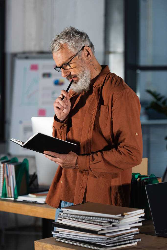 přemýšlivý šedovlasý podnikatel v brýlích držící pero a dívající se do zápisníku v kanceláři - Fotografie, Obrázek