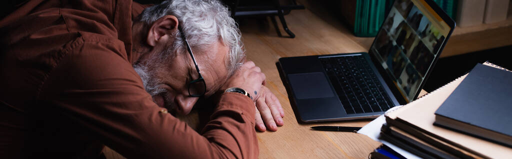 uupunut keski-ikäinen liikemies nukkumassa lähellä kannettavaa tietokonetta ja muistikirjoja toimistossa yöllä, banneri - Valokuva, kuva