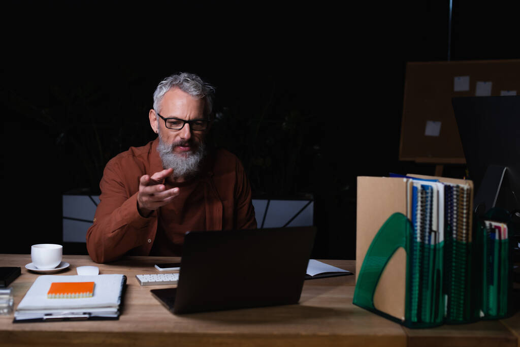 Gözlüklü, sakallı, orta yaşlı bir adam bilgisayardaki görüntülü sohbet sırasında gece bilgisayarını işaret ediyor. - Fotoğraf, Görsel
