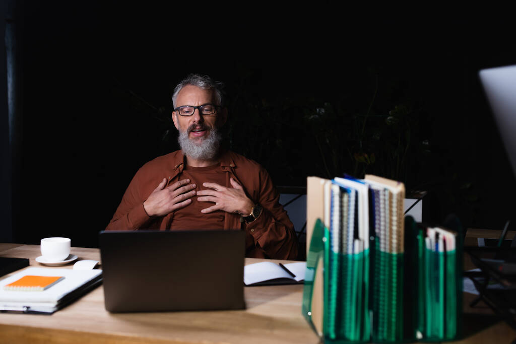 homme d'affaires satisfait toucher la poitrine et en regardant ordinateur portable près tasse de café et ordinateurs portables dans le bureau sombre - Photo, image