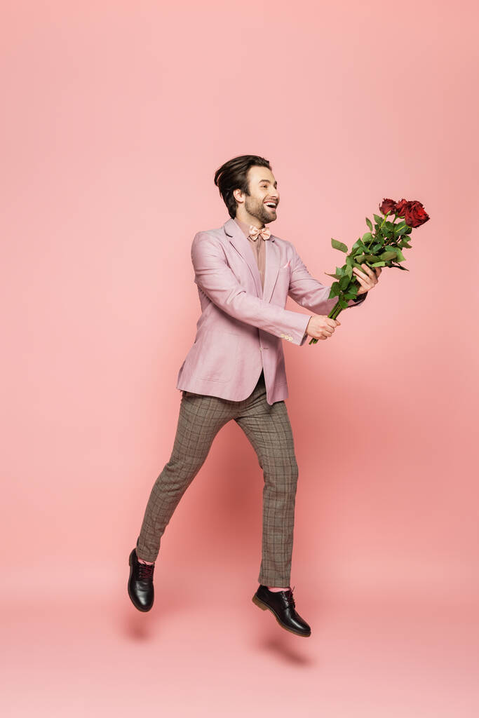 Volledige lengte van positieve gastheer van evenement met rozen tijdens het springen op roze achtergrond  - Foto, afbeelding