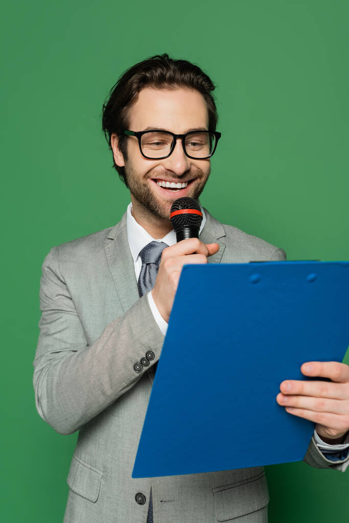 Frohe Botschaft Anker in Anzug und Brille sprechen in Mikrofon, während Klemmbrett isoliert auf grün halten  - Foto, Bild