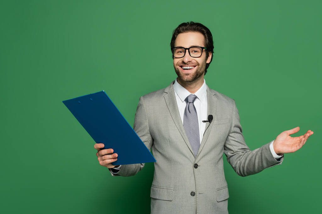 Ведущий новостей в костюме и очках держит планшет, жестикулируя на зеленом фоне  - Фото, изображение