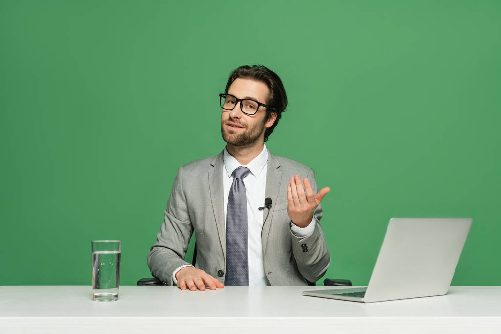 bearded news anchor in eyeglasses and suit talking near laptop isolated on green - Photo, Image