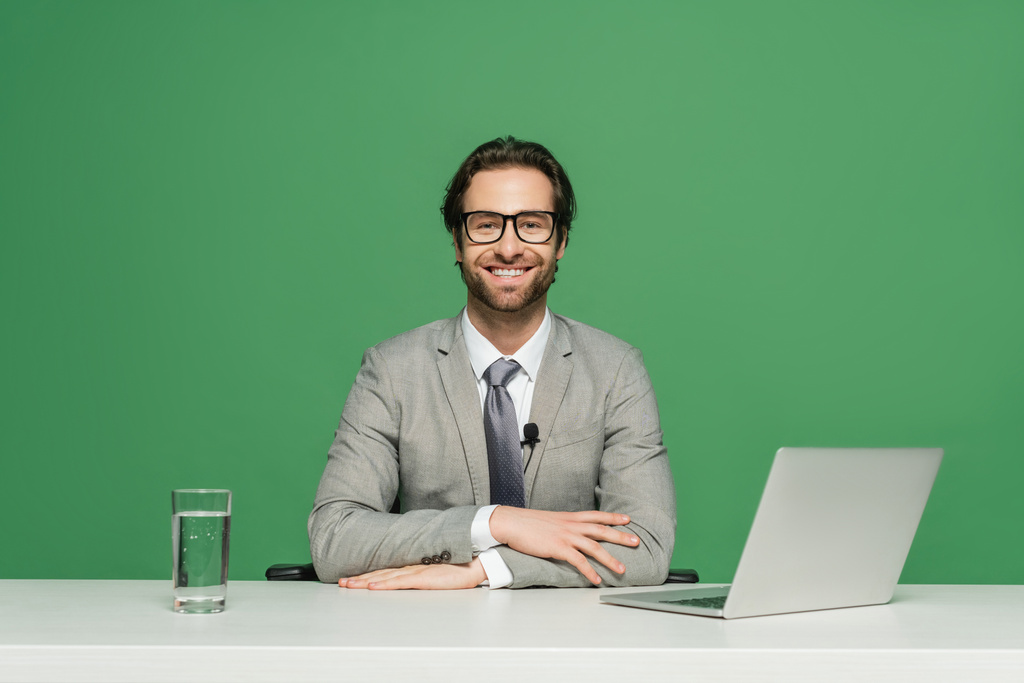 presentador de noticias barbudo en gafas y traje sonriendo mientras está sentado cerca de la computadora portátil aislado en verde - Foto, Imagen