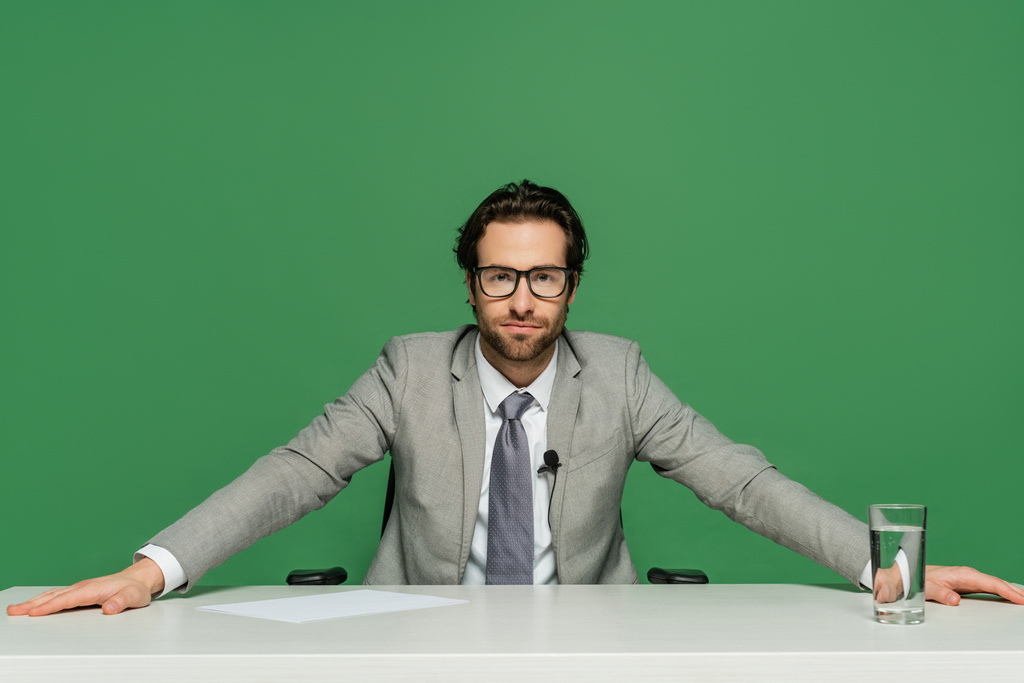 ραδιοτηλεοπτικός οργανισμός με γυαλιά ηλίου και γκρι κοστούμι που κάθεται στο γραφείο απομονωμένο σε πράσινο - Φωτογραφία, εικόνα