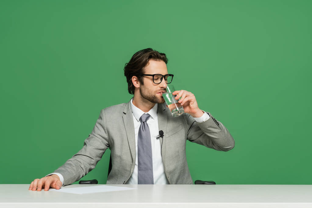 broadcaster in eyeglasses sitting at desk and drinking water isolated on green - Photo, Image