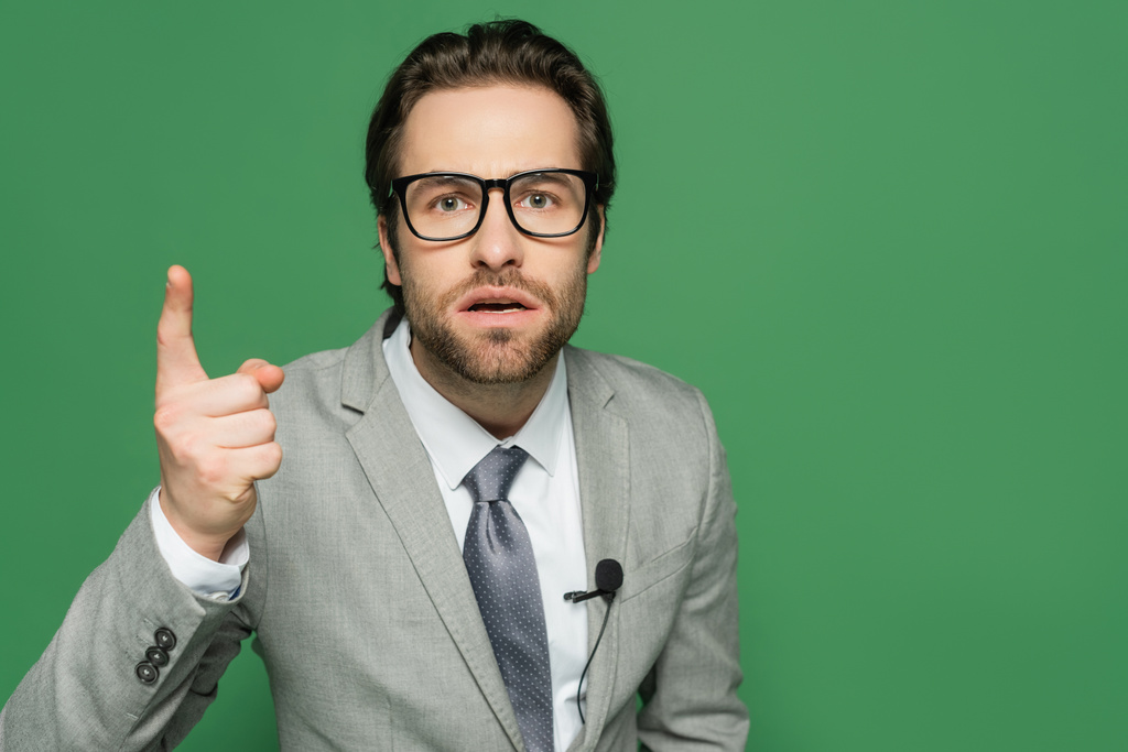 Эмоциональный ведущий новостей в очках и костюмах, указывающий пальцем на зеленый  - Фото, изображение