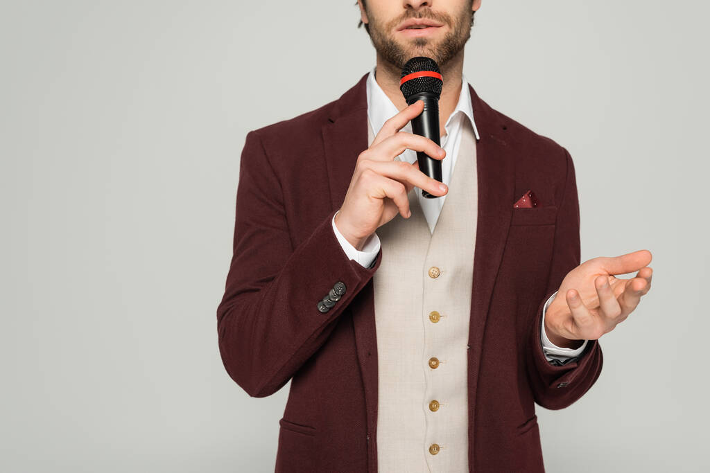 Ausgeschnittene Ansicht eines bärtigen Moderators in formeller Kleidung, der ein Mikrofon in der Hand hält und isoliert auf grau spricht  - Foto, Bild