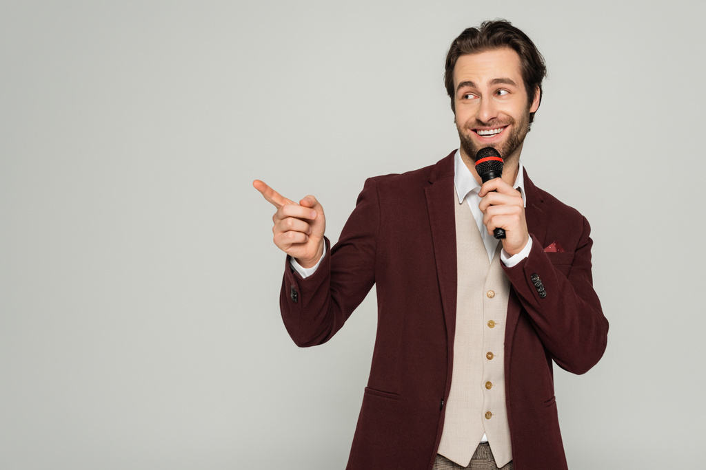 позитивний шоумен у формальному носінні, що розмовляє мікрофоном і вказує пальцем ізольовано на сірому
  - Фото, зображення
