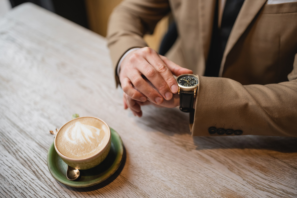 ラテアートでカプチーノのカップの近くに腕時計に触れる男のクロップドビュー  - 写真・画像