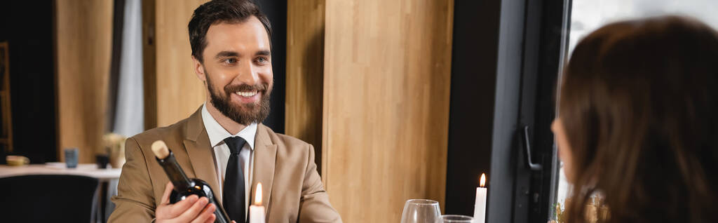 homme barbu souriant tout en tenant la bouteille avec du vin près de petite amie pendant le dîner festif le jour de la Saint-Valentin, bannière  - Photo, image