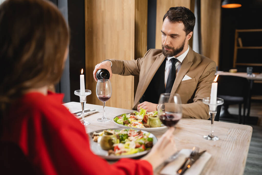 bearded man pouring red wine in glass near girlfriend during dinner on valentines day - Photo, Image