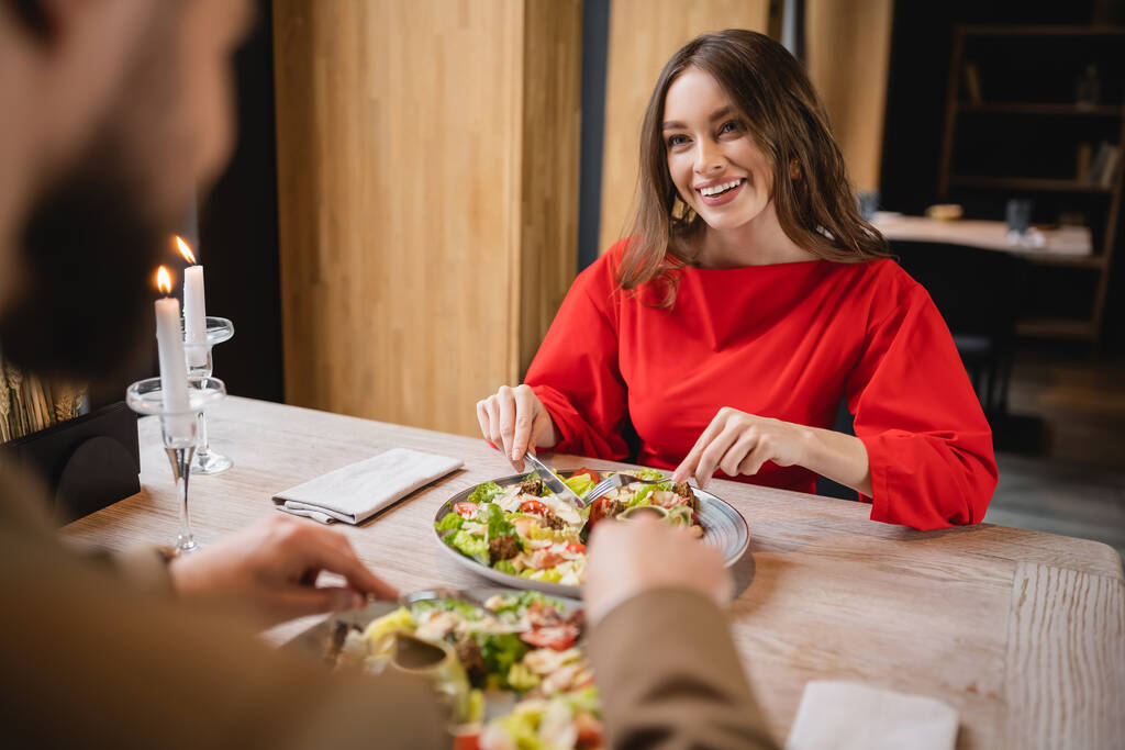 mujer alegre sonriendo y mirando al novio mientras sostiene cubiertos cerca de la ensalada en el restaurante en el día de San Valentín - Foto, Imagen
