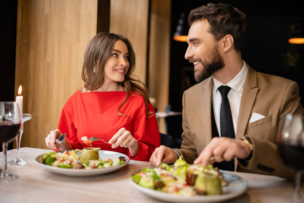 veselý mladý pár ve svátečním oblečení při pohledu na sebe při jídle během oslav na Valentýna  - Fotografie, Obrázek