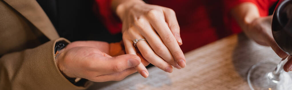 καλλιεργημένη άποψη του άνδρα που κρατά το χέρι της γυναίκας με δαχτυλίδι αρραβώνων στο δάχτυλο την ημέρα του Αγίου Βαλεντίνου, πανό  - Φωτογραφία, εικόνα