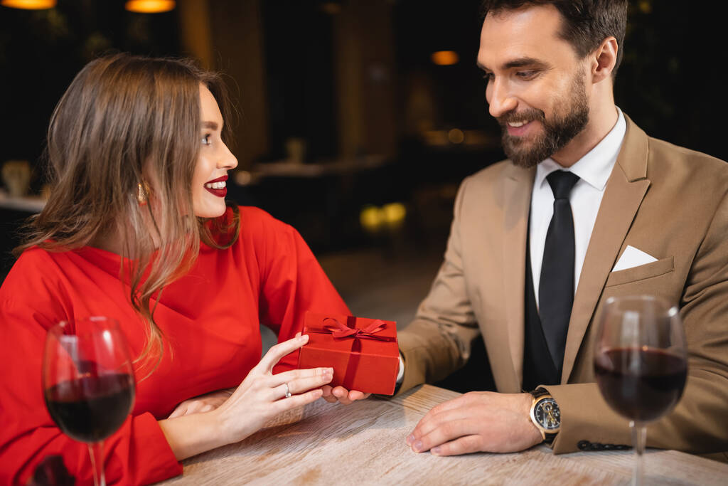 szczęśliwy mężczyzna trzyma prezent w pobliżu radosnej kobiety w Walentynki  - Zdjęcie, obraz