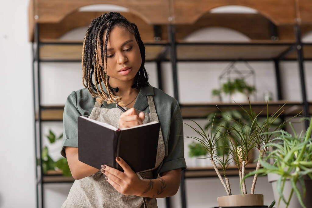 アフリカ系アメリカ人の若い花屋さん花の店の緑の植物の近くのノートブックで注文書を書く - 写真・画像