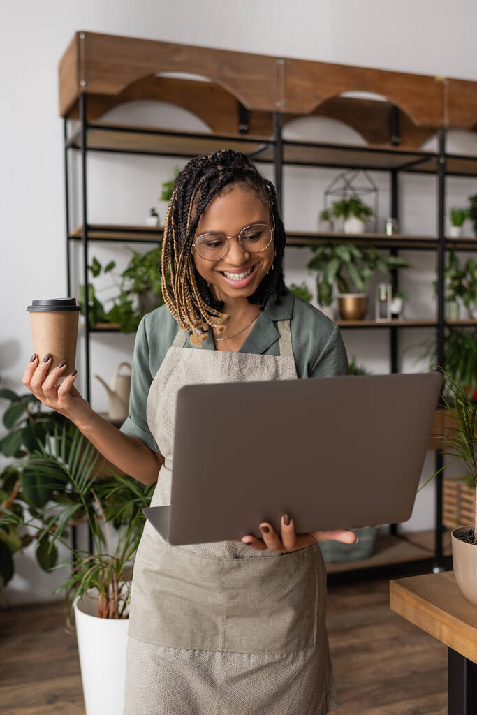 Afrikanisch-amerikanischer Blumenhändler mit Imbissgetränk lächelt während Videoanruf auf Laptop im Blumenladen - Foto, Bild