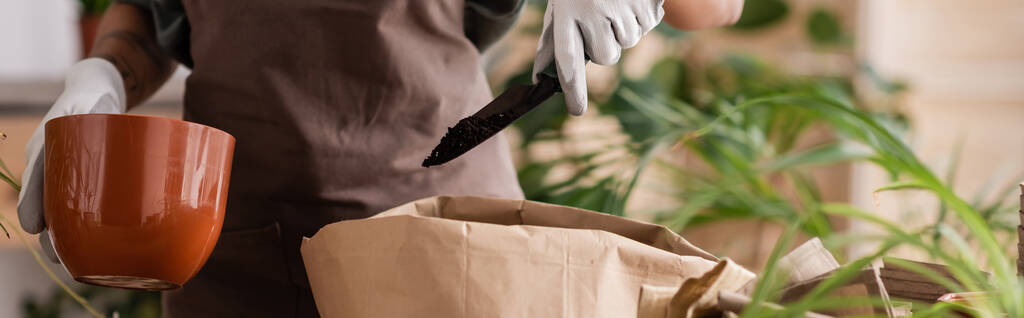 vista cortada de florista americano africano segurando vaso perto de saco de papel e colher de jardim com solo perto de plantas desfocadas, bandeira - Foto, Imagem