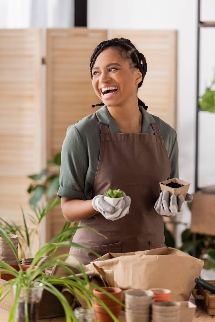 Αφροαμερικανός ανθοπώλης με φυτό και γλάστρα να γελάει κοιτώντας αλλού στο ανθοπωλείο - Φωτογραφία, εικόνα