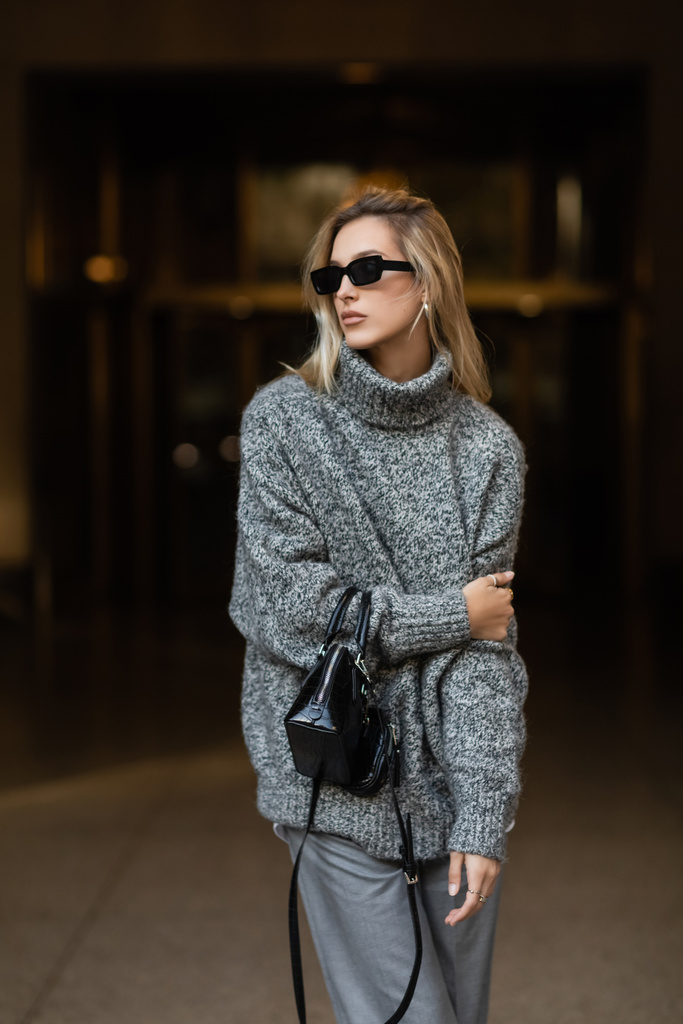 Stylische Frau mit Sonnenbrille und grauem Pullover steht mit schwarzer Handtasche in New York  - Foto, Bild